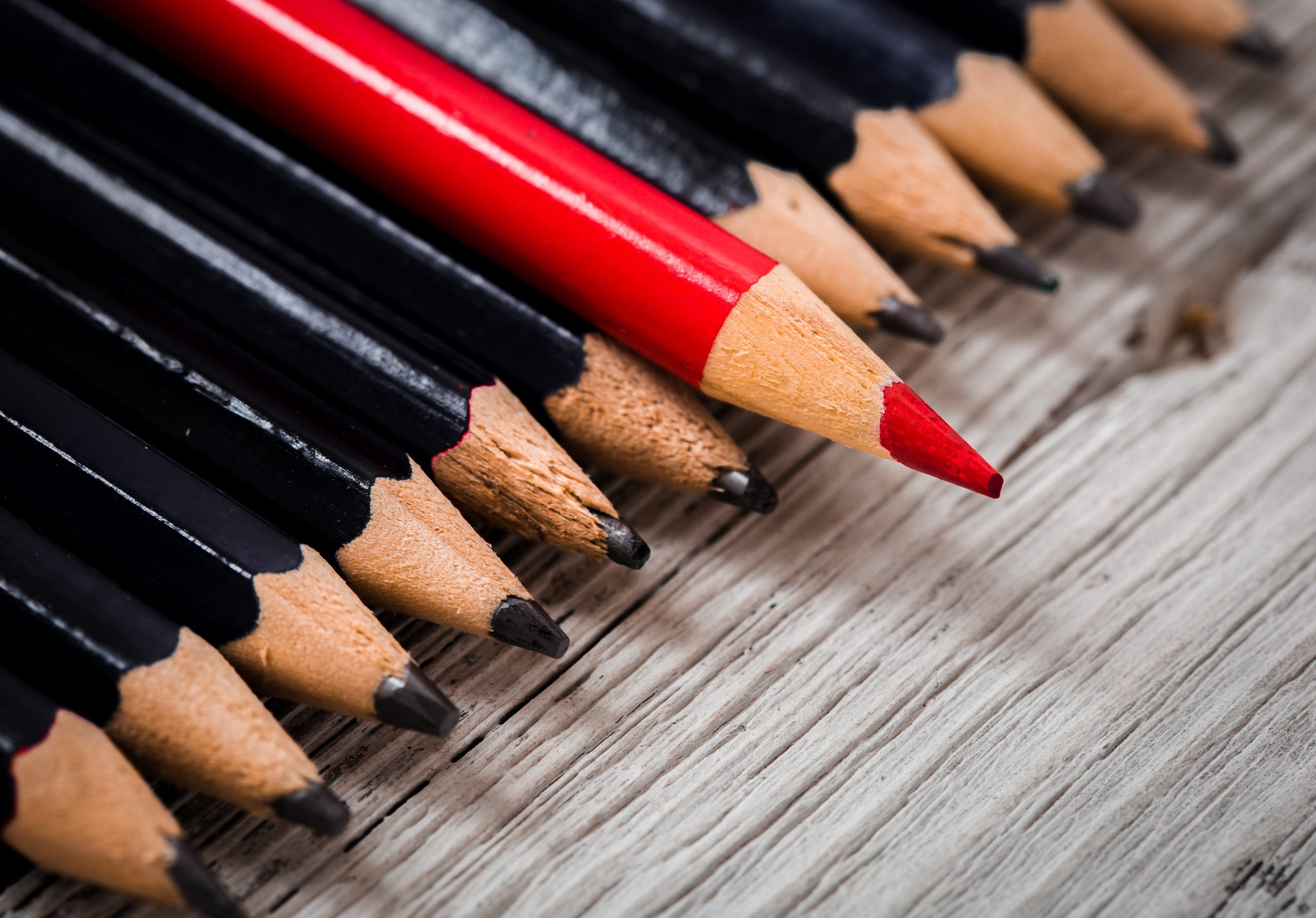 Red pencil stands out from the crowd of black  on a wooden white background.